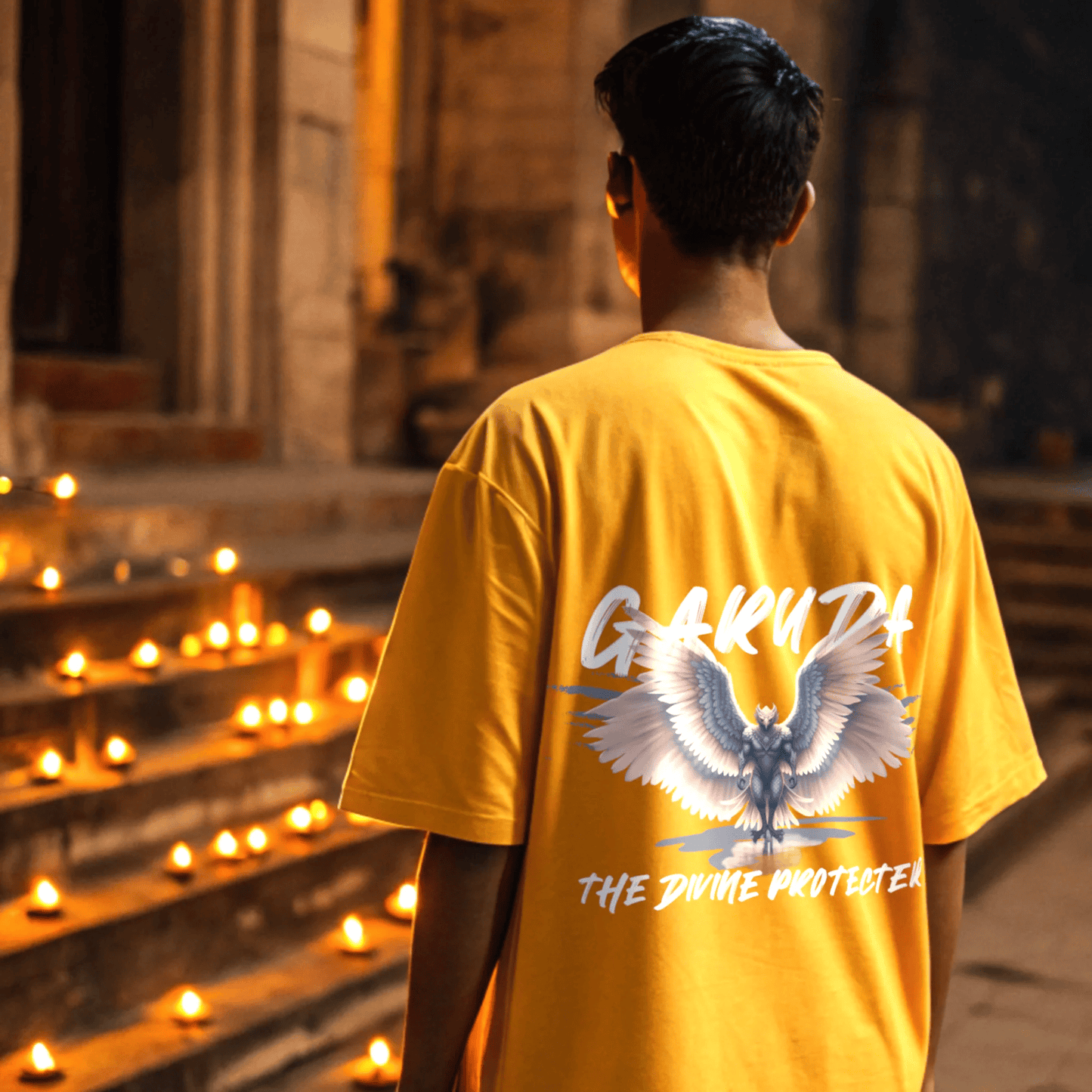 Person wearing a golden yellow t-shirt with a graphic design and garuda the divine protector text, standing in front of lit candles By OFF THE RACK