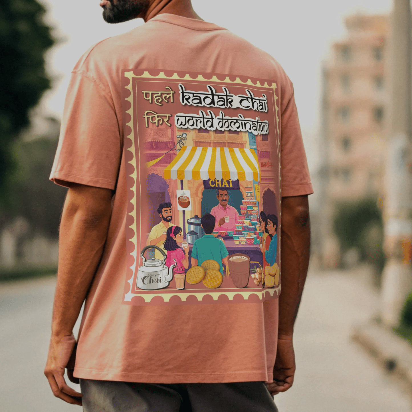male model wearing a coral t-shirt with a colorful graphic of KADAK CHAI WORLD DOMINATION on the back, standing outdoors by OFF THE RACK