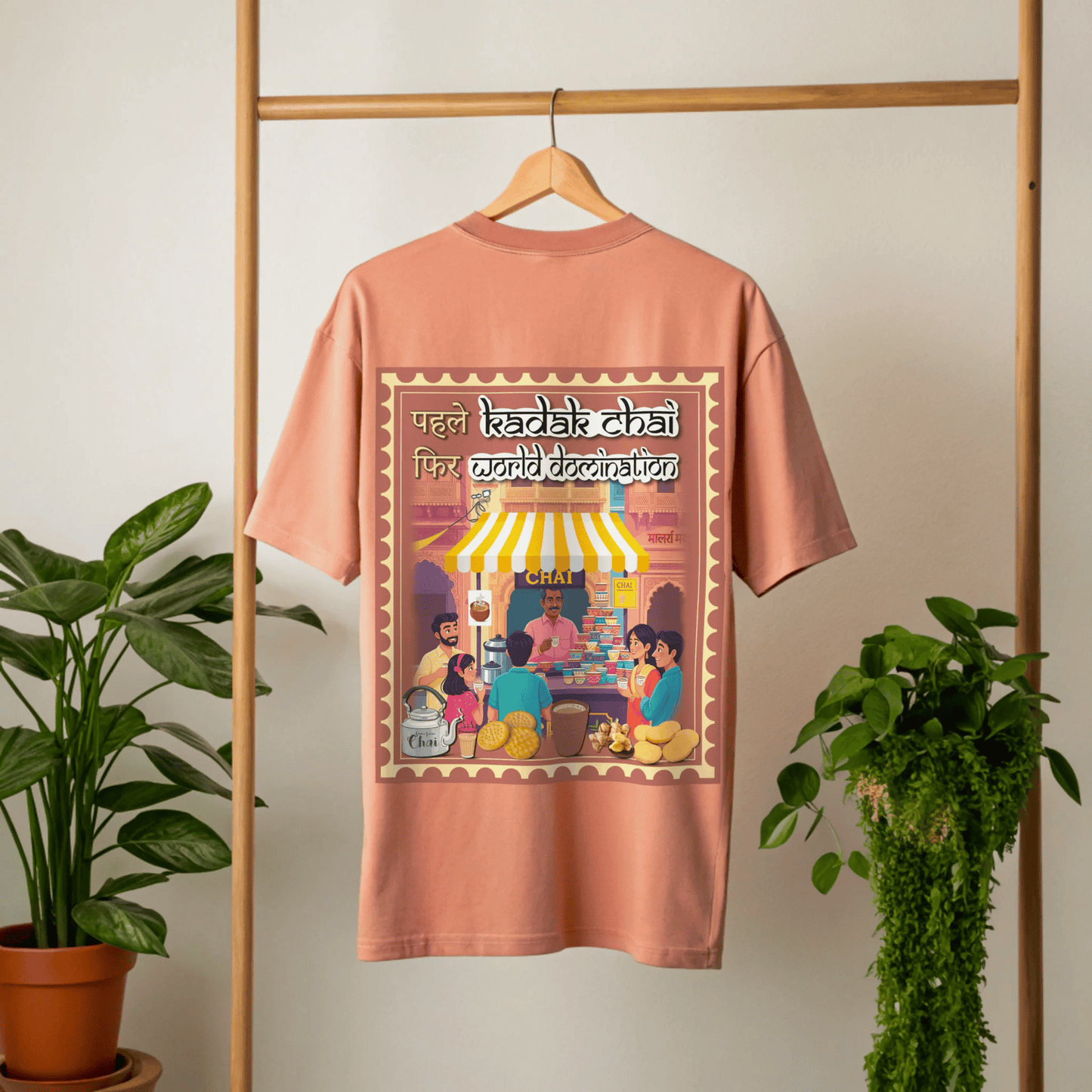 coral t-shirt with a graphic design of people around a chai stall on a hanger against a white wall with plants by the side By OFF THE RACK.