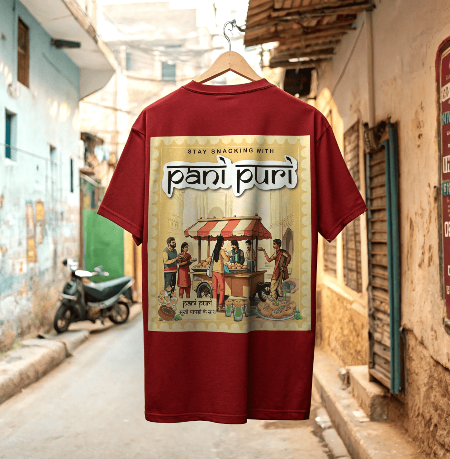 Red t-shirt with 'pani puri' graphic design on a hanger against a desi street background By OFF THE RACK