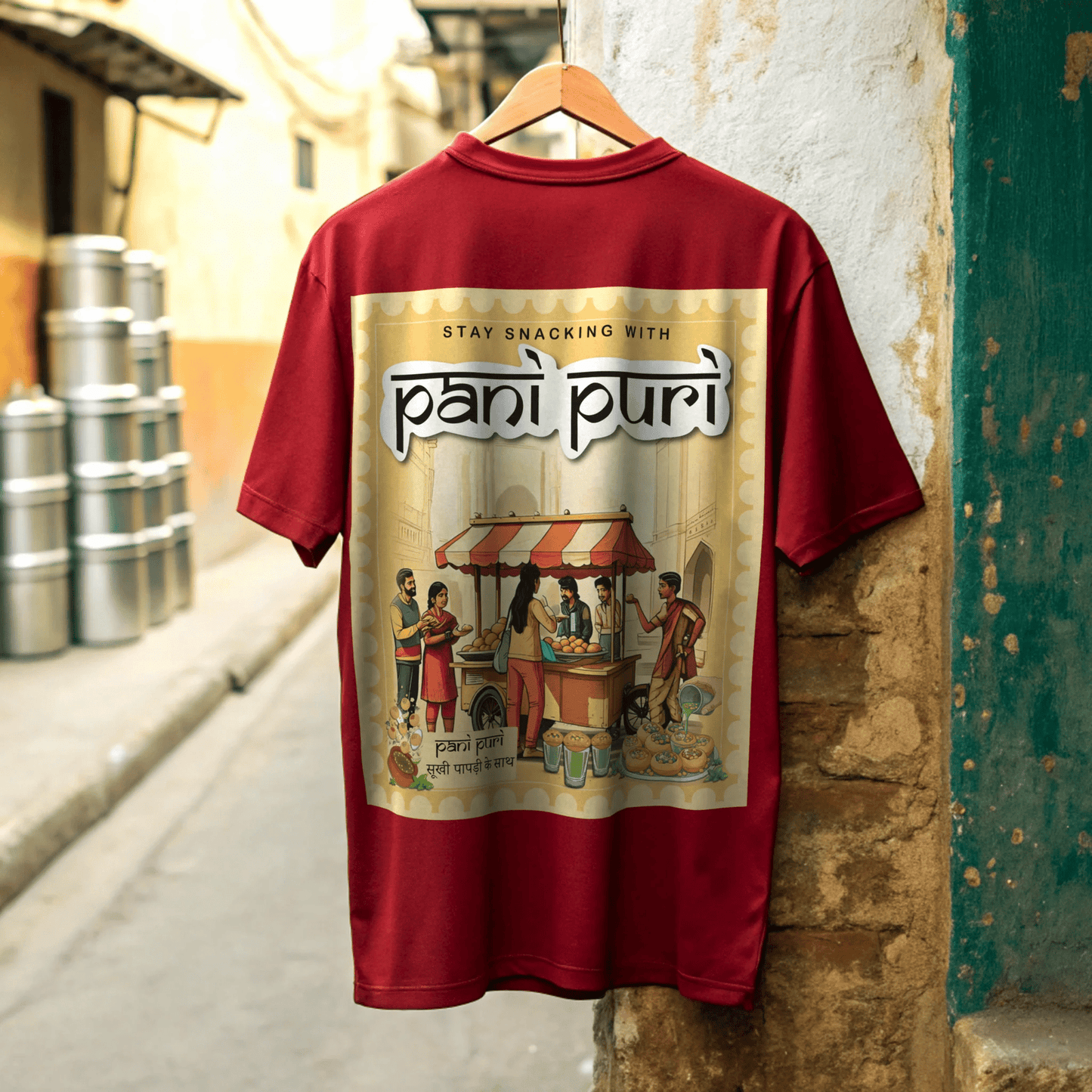 Red t-shirt with 'pani puri' graphic design hanging on a wooden hanger against a textured wall in small town street by OFF THE RACK
