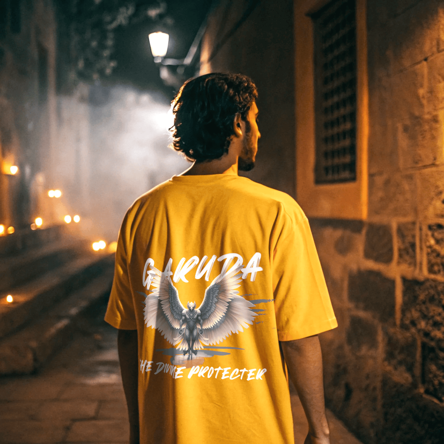 Person wearing a golden yellow t-shirt with 'Garuda' design and GARUDA THE DIVINE PROTECTOR text, standing in a dimly lit street by OFF THE RACK.