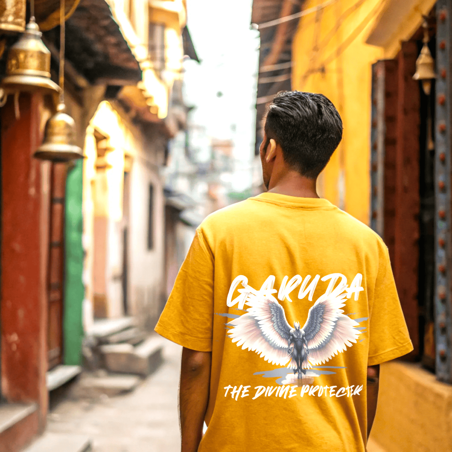Person wearing a golde yellow t-shirt with garuda the divine protector text and graphic in an alleyway by OFF THE RACK.