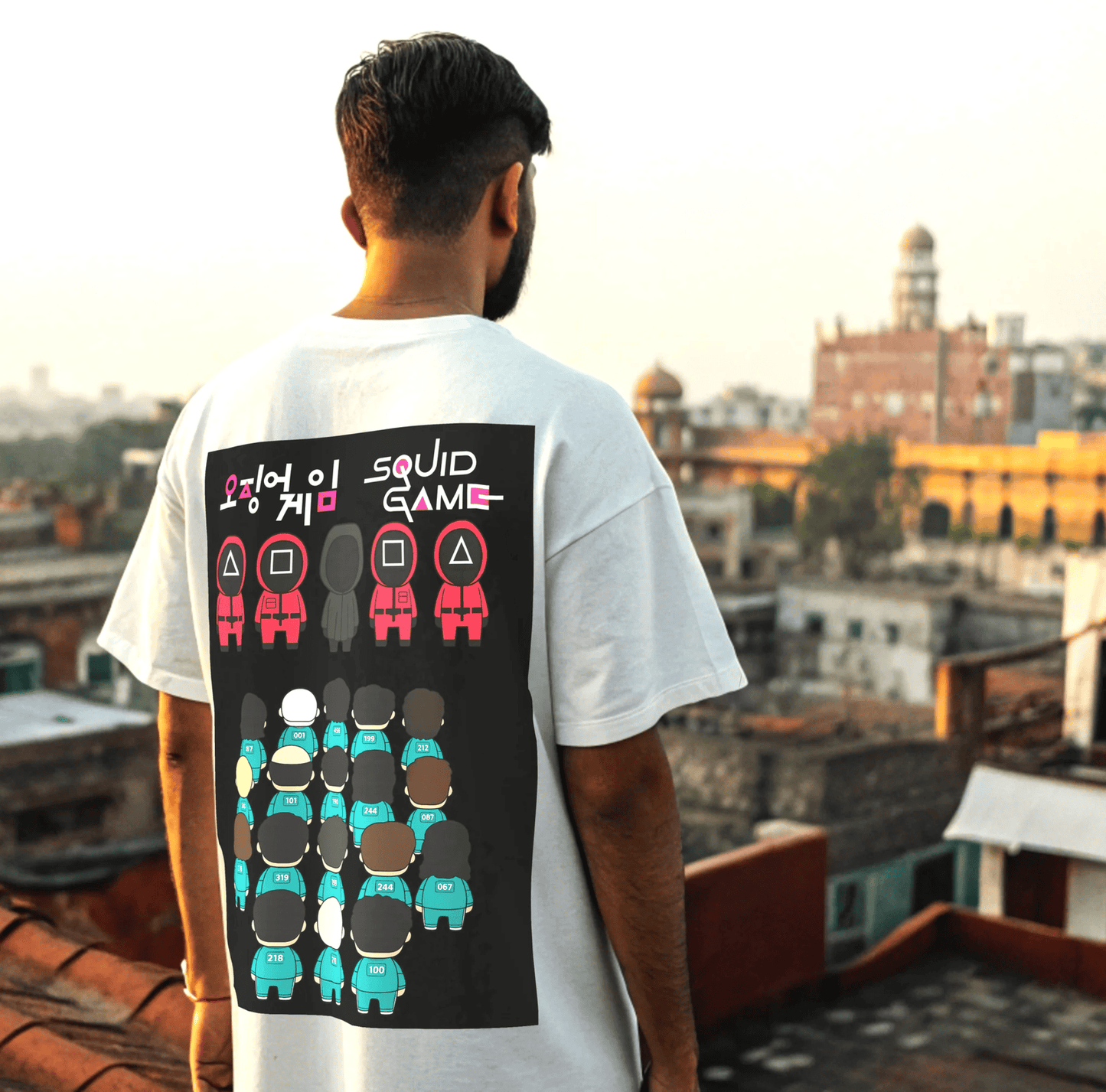 Person wearing a white t-shirt with a 'Squid Game' design on the back, standing on a rooftop with a cityscape in the background By OFF THE RACK.