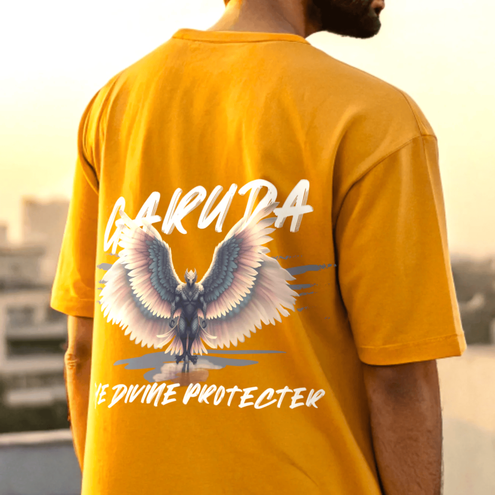 Person wearing a golden yellow t-shirt with garuda design and garuda the divine protector text on the back, standing outdoors by OFF THE RACK