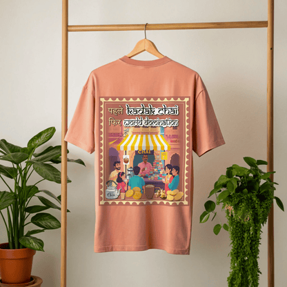 coral t-shirt with a graphic design of people around a chai stall on a hanger against a white wall with plants by the side By OFF THE RACK.