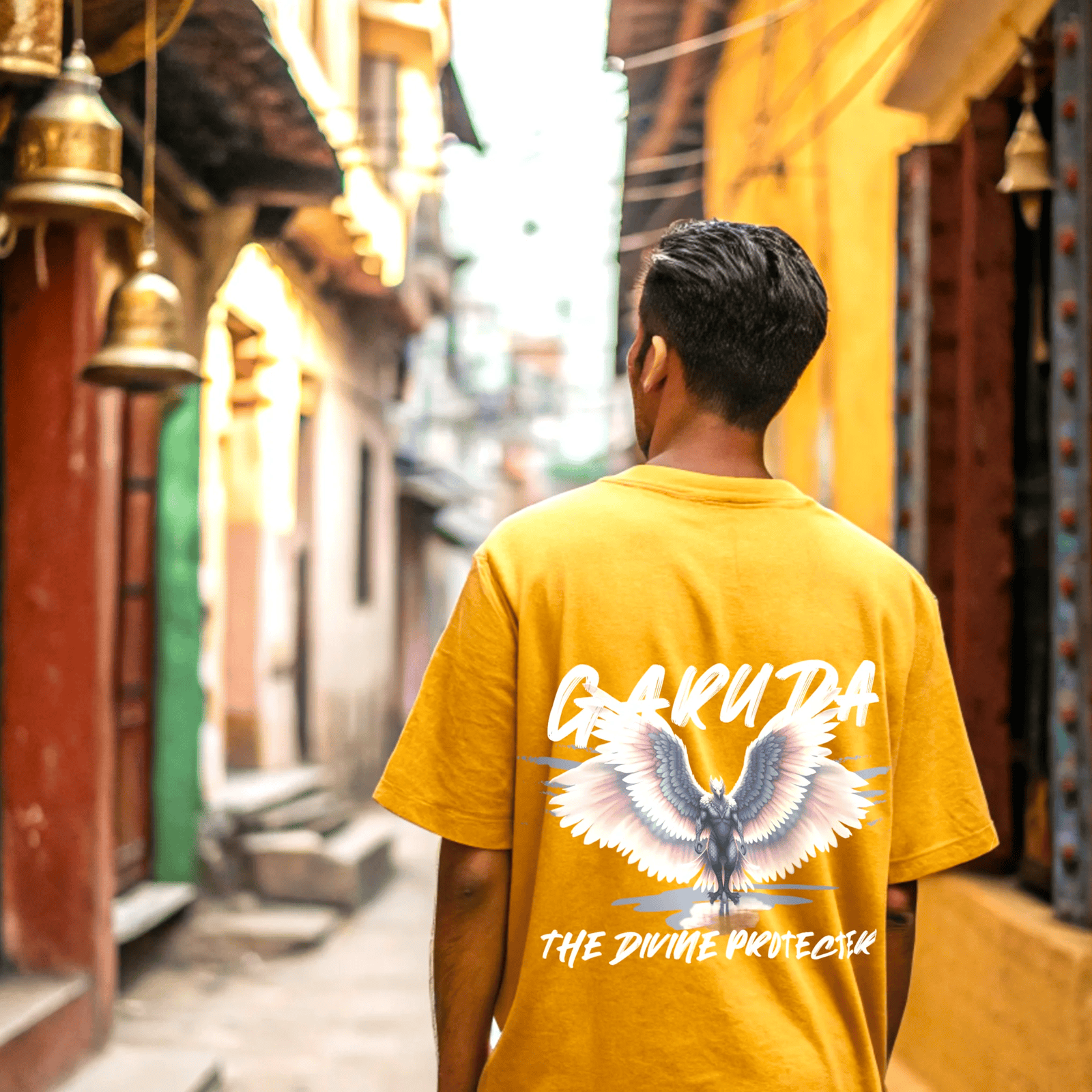 Person wearing a golde yellow t-shirt with garuda the divine protector text and graphic in an alleyway by OFF THE RACK.